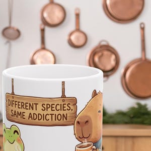 Puede incluir: Taza de cerámica blanca con un dibujo de una rana y un carpincho tomando café o té. La taza tiene el texto "DIFFERENT SPECIES, SAME ADDICTION" en un letrero de madera. En el fondo, hay sartenes de cobre colgadas.