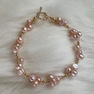 Può includere: Un delicato braccialetto con perle d'acqua dolce color rosa blush infilate su filo dorato. Il braccialetto ha una chiusura a toggle e piccole perline dorate. Le perle hanno una forma naturale e irregolare.