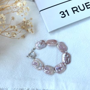 May include: A bracelet featuring large, irregularly shaped, light pink pearls. The pearls are strung together with small white beads and a silver toggle clasp. The bracelet is displayed on a white, textured surface.