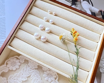 Pode incluir: Uma caixa de joias de madeira com interior creme contém vários brincos de pérolas brancas de tamanhos variados. Um raminho de flores amarelas e brancas adiciona um toque de cor. Renda branca delicada é visível.
