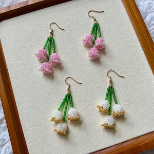 May include: Four pairs of beaded dangle earrings. Two pairs feature pink beaded flower clusters with green stems and gold-colored hooks. The other two pairs have white beaded flower clusters with gold-colored accents and green stems.