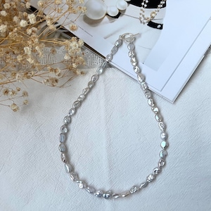 Pode incluir: Um colar prateado com pérolas irregulares cinza claro. O colar, com fecho de alternância, é exibido em uma superfície branca. As pérolas têm um brilho lustroso, refletindo a luz. Aproximadamente 40 cm de comprimento.