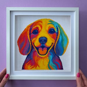 May include: A square white frame containing a cross-stitch embroidery of a dog's face. The dog is rendered in vibrant, multi-coloured hues including yellow, orange, red, blue, and purple, featuring a wide smile with a visible tongue.