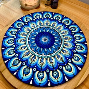 May include: A round, hand-painted mandala in shades of blue, teal, and white. The intricate design features concentric circles with dot patterns and eye-like shapes. The artwork is displayed on a wooden surface.