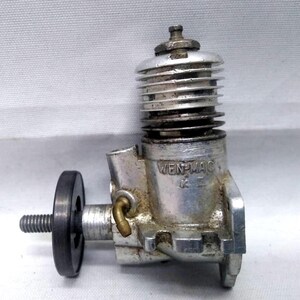 May include: A vintage silver-toned model airplane engine with the text "WEN-MAC" visible. The engine features a ribbed cylinder head, a black flywheel, and a threaded shaft. The engine is designed for model aircraft.