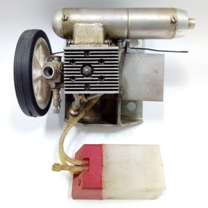 May include: A vintage model engine with a silver-colored body, a black tire, and a white wheel. The engine has a ribbed cylinder head and a fuel tank with a red top. The engine is designed for model airplanes or cars.