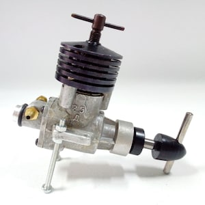 Puede incluir: Un pequeño motor plateado con aletas de refrigeración negras y una hélice negra. El motor tiene un botón de ajuste marrón y el número "2.5" es visible. Diseñado para modelos de aviones o aplicaciones similares.