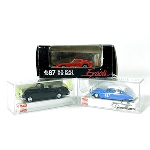 May include: Three miniature model cars in clear display cases. A red sports car is in a black box. The other two are a black convertible and a blue sedan. The models are 1:87 scale.