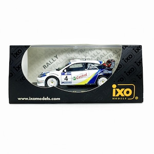 May include: A model of a Ford Fiesta rally car, white with blue and yellow accents, number 4, presented in a black box. The car has sponsor logos, including "Castrol". The box displays the "ixo models" logo and the website address www.ixomodels.com.