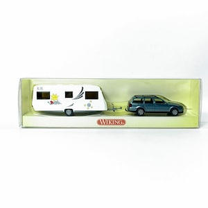 以下が含まれることがあります： 白いおもちゃのキャンピングカーと青いステーションワゴンがセットになったおもちゃ。太陽のグラフィックが描かれています。透明なプラスチックケースに入っています。「Wiking」のブランド名が見えます。