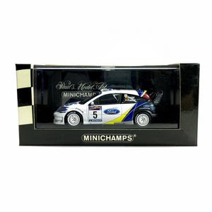 May include: A white, blue, and yellow Ford Focus rally car model in a black display case. The model car has the number 5 on the side and the Ford logo. The display case has the text "Paul's Model Art" and "MINICHAMPS".