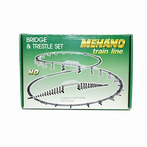 Può includere: Una scatola verde con un set di treni con il testo "Bridge & Trestle Set" e "MeHano train line". La scatola mostra un diagramma di un binario ferroviario con ponti e cavalletti. Il set include 14 paia di supporti.