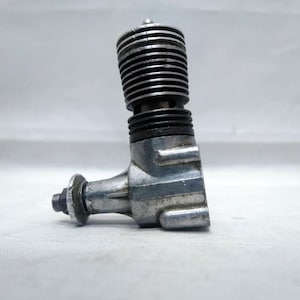 May include: A silver-colored model engine with a ribbed cylinder head and a black rubber gasket. The engine has a small, circular flywheel and is designed for model aircraft or other small-scale applications.