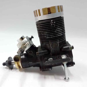 May include: A small, black internal combustion engine with a silver and gold-colored components. The engine has a ribbed cylinder and a silver top with gold-colored stripes. The engine is designed for model aircraft or similar applications.