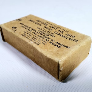 Puede incluir: Un botiquín de primeros auxilios rectangular vintage en una caja de cartón marrón. La caja está etiquetada como "INDIVIDUAL TROOP CAMOUFLAGED, DRESSING, FIRST AID, FIELD." Las dimensiones son 10 x 18 cm. La caja está ligeramente desgastada.
