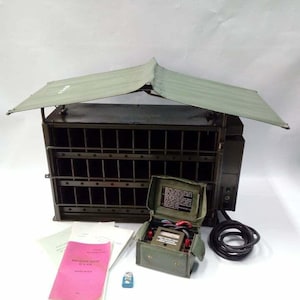 Peut inclure: Un ensemble de radio de campagne militaire vert olive avec un auvent protecteur. L'ensemble comprend une petite bo&icirc;te ouverte avec des jauges, un c&acirc;ble noir enroul&eacute;, un manuel rose et un petit objet bleu. La radio a une fa&ccedil;ade en forme de grille.