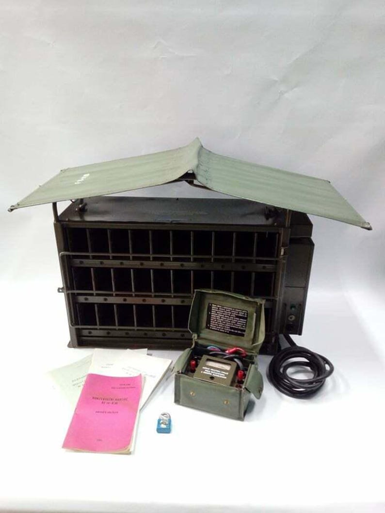 Peut inclure: Un ensemble de radio de campagne militaire vert olive avec un auvent protecteur. L'ensemble comprend une petite bo&icirc;te ouverte avec des jauges, un c&acirc;ble noir enroul&eacute;, un manuel rose et un petit objet bleu. La radio a une fa&ccedil;ade en forme de grille.