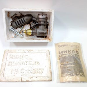 May include: A vintage model engine kit, comprising engine components, tools, and packaging. The engine is silver and gold-toned, with a black exhaust. The packaging includes a white foam container and a paper packet with Cyrillic text.