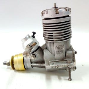 May include: A silver and gold model engine with the text "MAC 65KY" visible. The engine has a cylindrical body with cooling fins, a carburetor, and a spark plug. The engine is designed for model aircraft or other small applications.