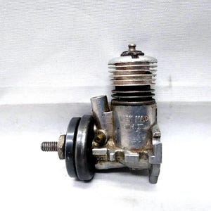 May include: A vintage silver-colored model engine with a cylindrical top and a side exhaust port. The engine has a black flywheel and a threaded bolt. The text "WEY MAC" is visible on the side.