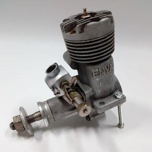 May include: A silver model engine, marked "ENYA (35) MODEL 2015". The engine features a cylindrical top with cooling fins, a carburetor, and a propeller shaft. It is designed for model aircraft or similar applications, with metric measurements.