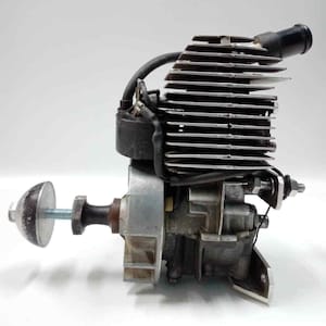 May include: A small, silver and black internal combustion engine with a finned cylinder head. The engine has a spark plug wire, a carburetor, and a mounting bracket. A bolt with a round, grey cap is attached to the engine.