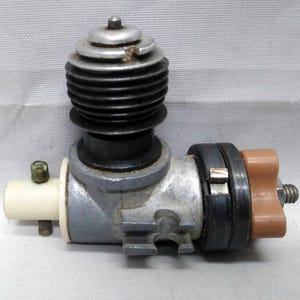 Puede incluir: Un motor de modelo vintage con cuerpo plateado, cilindro negro acanalado y un componente marrón. El motor tiene una pieza cilíndrica blanca y varios accesorios metálicos. Probablemente para modelos a escala.