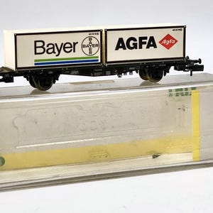 Könnte beinhalten: Ein Modell-Eisenbahnwagen mit den Logos Bayer und AGFA auf weißem Hintergrund. Der Wagen ist schwarz mit Rädern und wird in einer transparenten Kunststoffbox mit dem Aufdruck TRIX präsentiert.