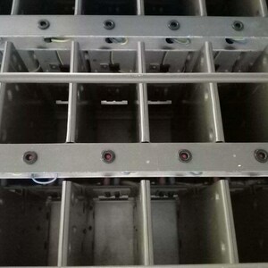 Peut inclure: Gros plan sur une structure m&eacute;tallique avec un design en forme de grille. La structure est compos&eacute;e de compartiments rectangulaires s&eacute;par&eacute;s par des barres m&eacute;talliques verticales et horizontales. De petits composants rouges et noirs sont visibles.