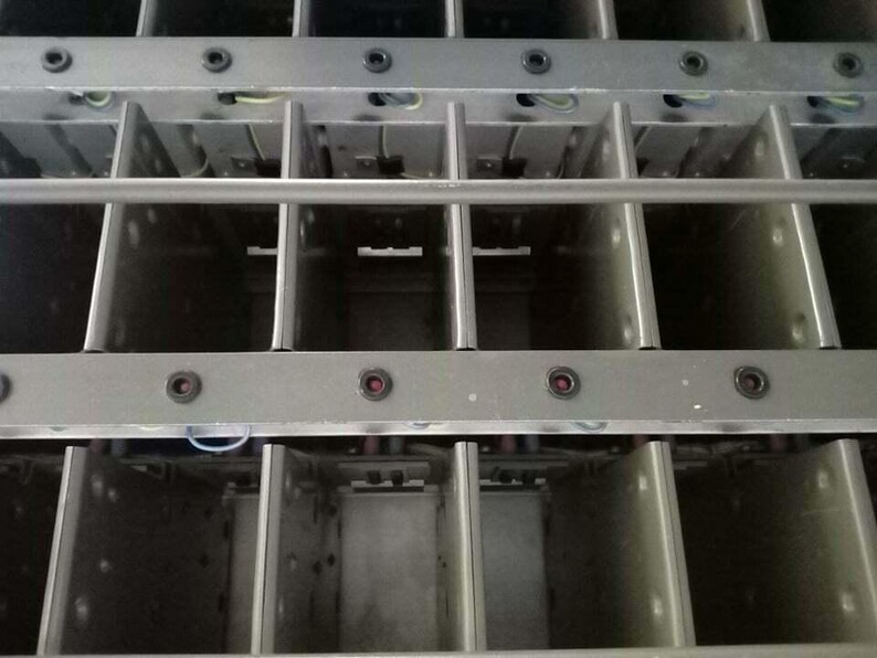 Peut inclure: Gros plan sur une structure m&eacute;tallique avec un design en forme de grille. La structure est compos&eacute;e de compartiments rectangulaires s&eacute;par&eacute;s par des barres m&eacute;talliques verticales et horizontales. De petits composants rouges et noirs sont visibles.