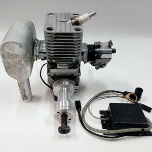 Puede incluir: Un pequeño motor de combustión interna de color plateado con bujía, escape y componentes adjuntos. El motor está diseñado para modelos de aviones o aplicaciones similares. Incluye una caja negra y cableado.