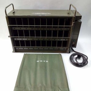 Peut inclure: Un appareil m&eacute;tallique vert olive avec une fa&ccedil;ade grillag&eacute;e et deux poign&eacute;es sur le dessus. Une housse assortie vert olive avec le texte "RF 10-K 30" est devant l'appareil. Un cordon d'alimentation noir est connect&eacute; &agrave; l'appareil.