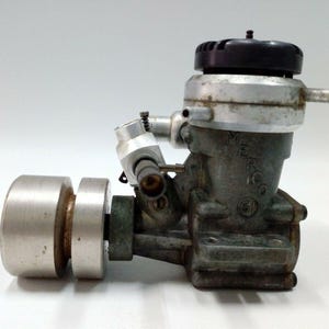 May include: A vintage model engine with a silver and black finish. The engine has a cylindrical flywheel and a complex arrangement of tubes and fittings. The body is a dark, aged metal. The engine is likely for a model airplane or car.
