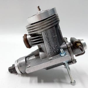 Puede incluir: Un motor de avión modelo plateado y gris vintage. El motor tiene un diseño cilíndrico con aletas de refrigeración, un carburador y una bujía. El texto "MAC 25XP" es visible en el lateral. El motor está montado sobre una base metálica.