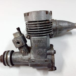 Puede incluir: Un motor de modelo Thunder Tiger 25 usado, hecho de metal plateado. El motor tiene un cuerpo cilíndrico con aletas de refrigeración, un carburador y un puerto de escape. El texto "THUNDER TIGER" y "25" son visibles.