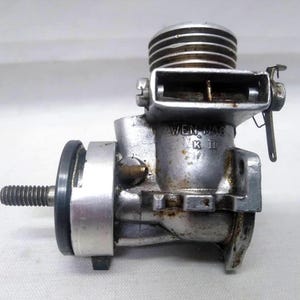 May include: A vintage silver-toned model engine with a ribbed cylinder head and a threaded shaft. The engine has the text "WEN MAC MK II" on its side. The engine is likely for a model airplane or car.