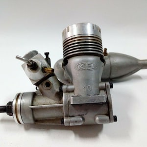 May include: A silver K&B 40 model engine, a small internal combustion engine, with a cylindrical cooling head and a muffler. The engine has the text "K&B" and "40" on its body. The engine is designed for model aircraft or other small-scale applications.