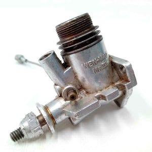 May include: A vintage silver-colored model engine with the text "WEN-MAC MK.III" visible. The engine has a threaded top, a small tube, and a bolt at the bottom. The engine is likely for a model airplane or car.