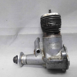 May include: A vintage model engine, primarily silver in color, with a cylindrical, ribbed top. The engine has a complex design with various mechanical components, including a circular base and a side-mounted element. The overall aesthetic suggests a focus on functionality and mechanical detail.