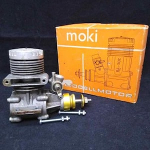 May include: A model engine with a silver body and a yellow and silver component, next to an orange box with the word "moki" and a diagram of an engine. The engine has a cylindrical top with horizontal ridges.