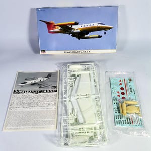 以下が含まれることがあります： U-36A Learjet J.M.S.D.F. の航空機モデルキット。 箱の絵は、青い空を背景に、黄色、白、赤のジェット機を示しています。 キットには、組み立てられていないプラスチック部品、デカール、および説明書が含まれています。