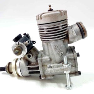 May include: A vintage silver-colored internal combustion engine with a cylindrical design. The engine has multiple cooling fins, a carburetor, and a spark plug. The letters "K/B" are visible on the engine block. A bolt is attached to the engine.
