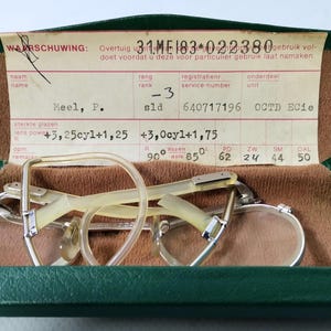 May include: A pair of vintage spectacles with a light-coloured frame and round lenses inside an open green case. The case has a brown interior and a printed paper label with text.