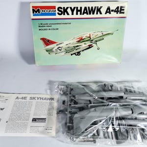 Peut inclure: Maquette Monogram Skyhawk A-4E. La boîte présente une illustration de l'avion en vol, avec des détails rouges et blancs. Le kit comprend des pièces en plastique non assemblées et des instructions.