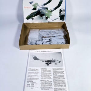 Puede incluir: Maqueta del avión OV-10A Bronco. La caja muestra una ilustración detallada del avión en camuflaje verde y gris. El kit incluye piezas de plástico sin montar, pintura e instrucciones.