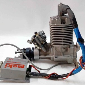 May include: A silver internal combustion engine with a ribbed cylinder head and attached carburetor. A grey box with the text "moki Ignition CE" is connected to the engine via wires and a grey cable. A blue and black cable is wrapped around the engine.