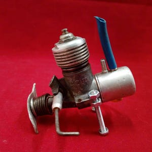 Può includere: Un motore modello vintage con corpo argentato, testata cilindrica alettata e tubo blu. Il motore è montato su una superficie rossa. La parola "CUR" è visibile sul motore.