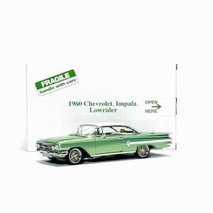May include: A metallic light green and white 1960 Chevrolet Impala lowrider model car with wire-spoke wheels. Behind the car is a white box with green text reading FRAGILE handle with care, 1960 Chevrolet Impala Lowrider, and OPEN HERE.