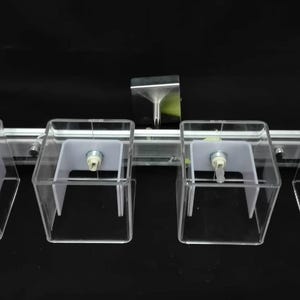 Op de afbeelding: Een heldere acryl display armatuur met vier vierkante compartimenten, elk met een wit interieur en een kleine metalen component. De armatuur is gemonteerd op een zilveren staaf met een reflecterend metalen stuk erboven.