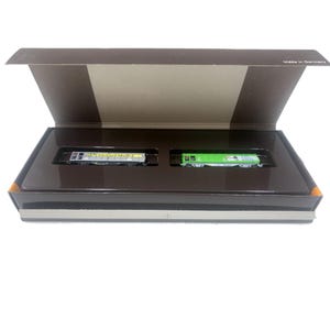 Puede incluir: Un juego en caja de dos vagones de tren en miniatura. Un vagón es plateado con texto amarillo y negro, el otro es verde brillante. La caja es marrón con un interior marrón oscuro y una tapa marrón claro. La caja está etiquetada como "Made in Germany."